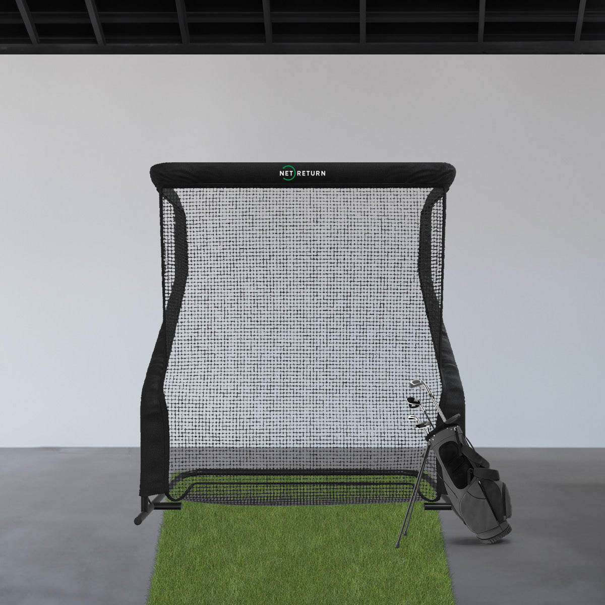 Golf practice net with metal frame stands on a green mat. A golf bag with clubs is leaning against it.